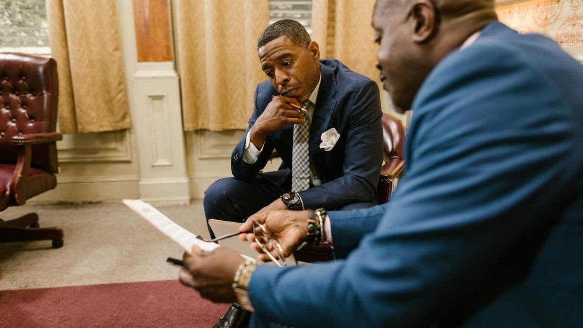 Two executives in business attire discuss documents in an office setting.