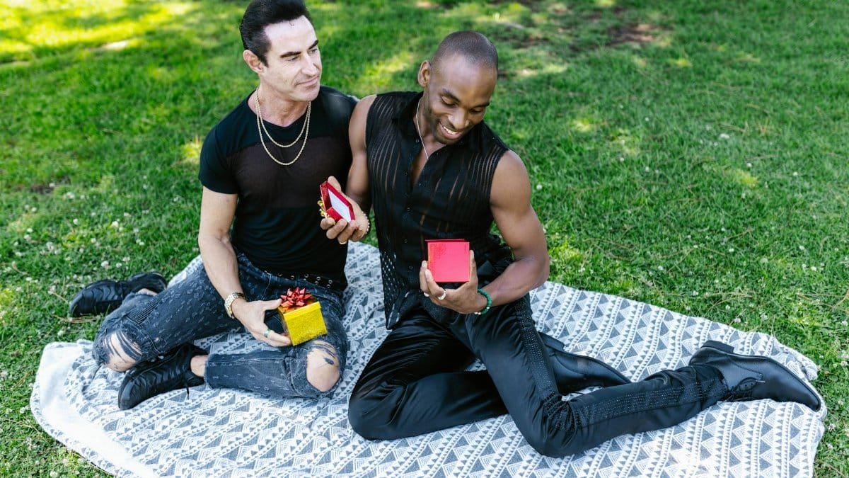 Two men sitting on a blanket in a park, exchanging gifts and smiling warmly.