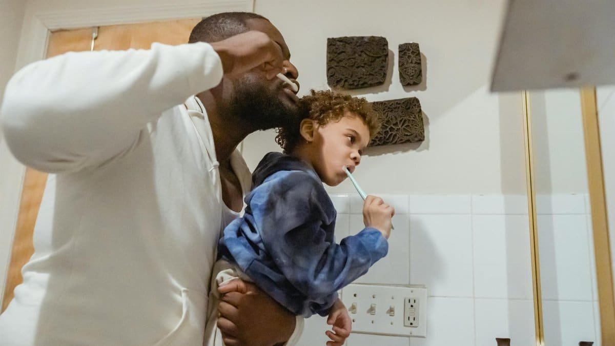 Side view of focused African American father with boy in hands brushing teeth with toothbrushes during morning routine in bathroom