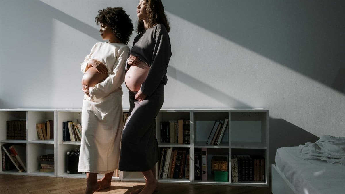 Two pregnant women embracing in a sunlit room, symbolizing love and anticipation.