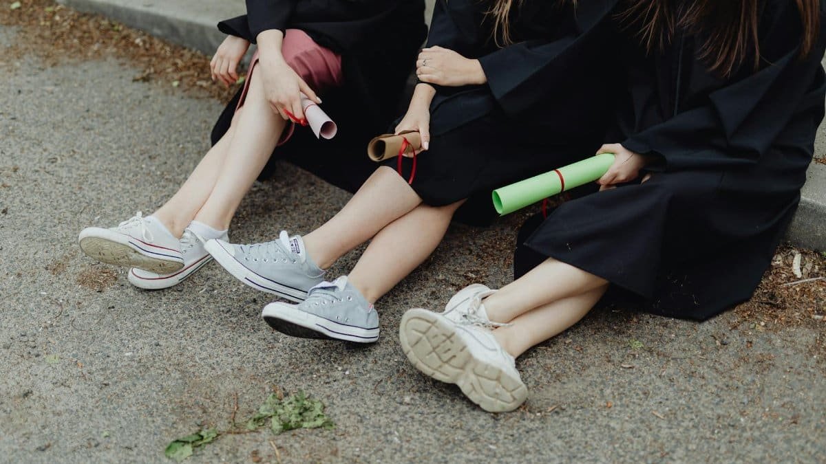 Three graduates in gowns sit on a roadside holding diplomas, legs crossed.