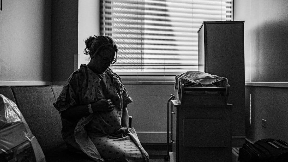 A thoughtful pregnant woman in a hospital room, captured in black and white.
