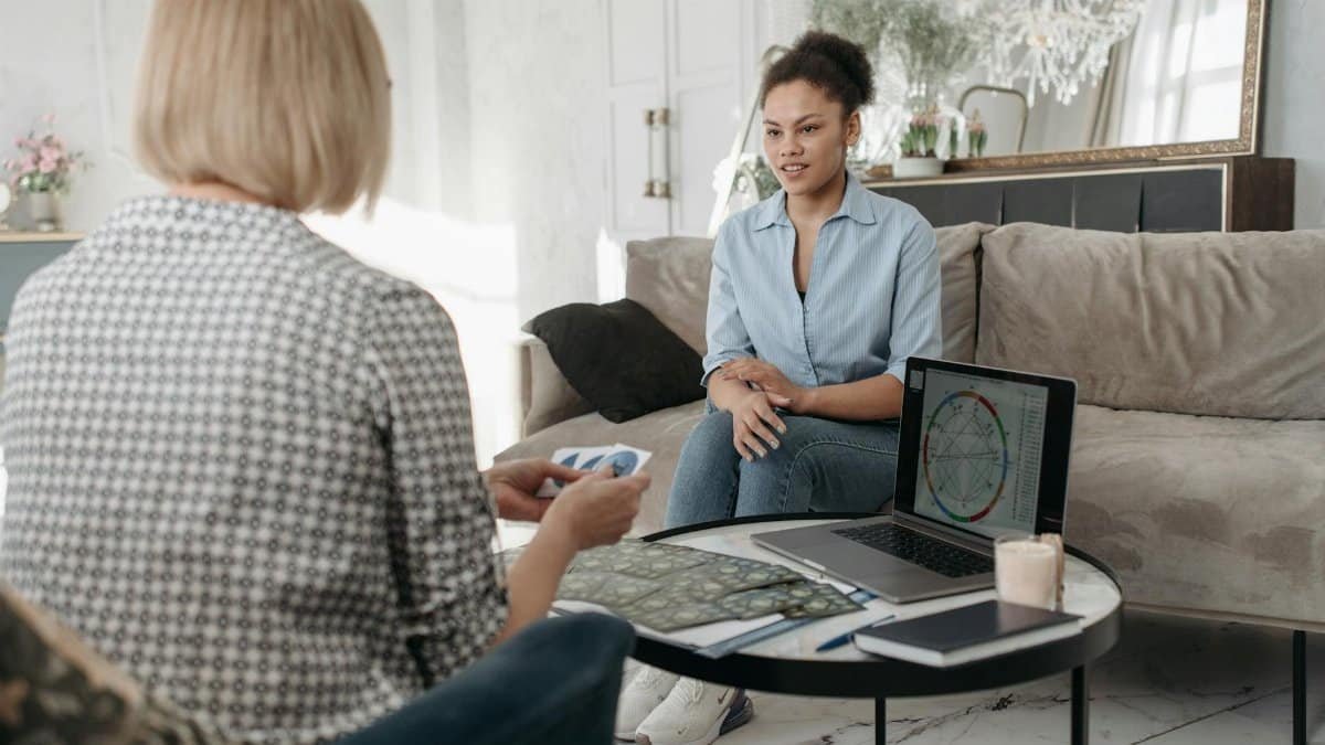 Women engaged in a tarot reading session with astrology charts on a laptop in a cozy living room.