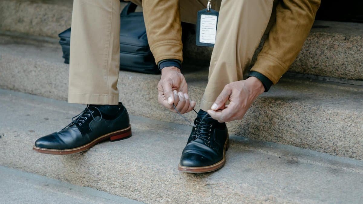Close-up of a man tying black leather shoes while sitting on stone steps outdoors.