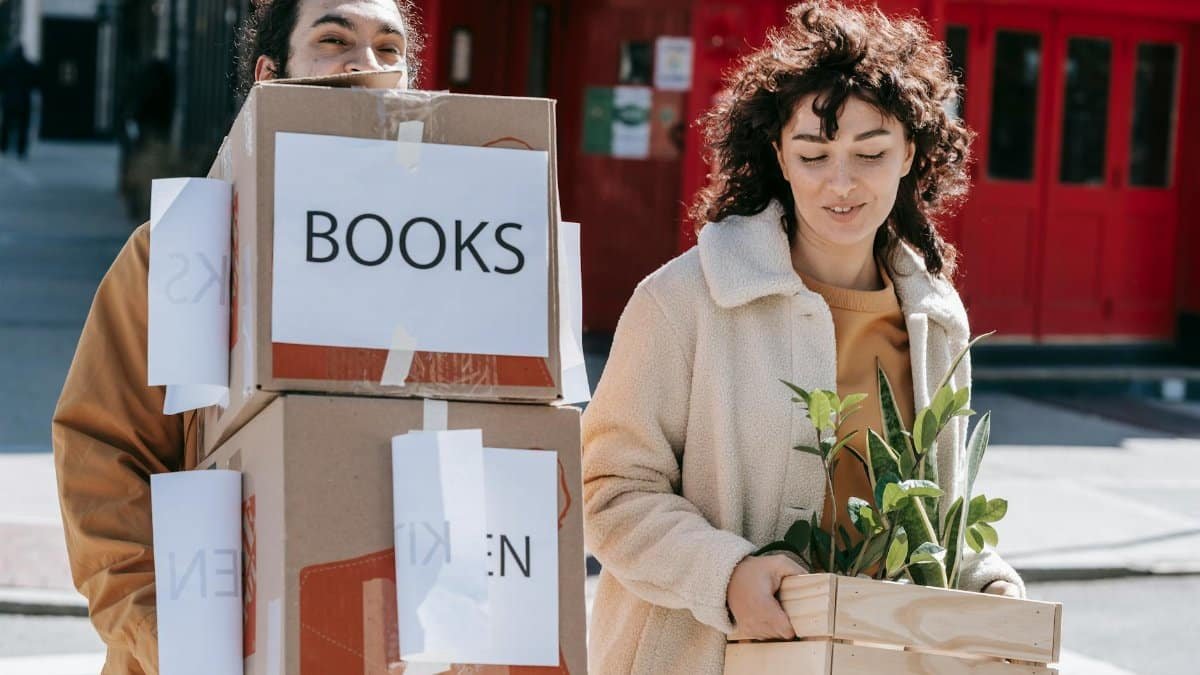 A joyful couple carrying boxes and plants while moving into a new home in an urban setting.