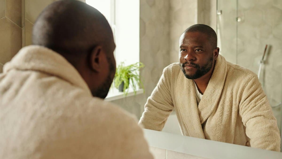 Adult man in a bathrobe looking at his reflection in a bathroom mirror. Morning routine concept.