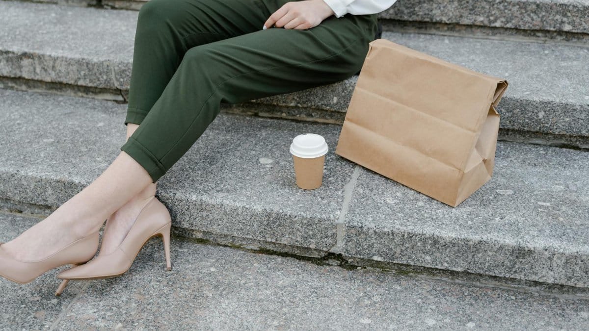 Woman sitting on steps with coffee and paper bag, stylish outfit.