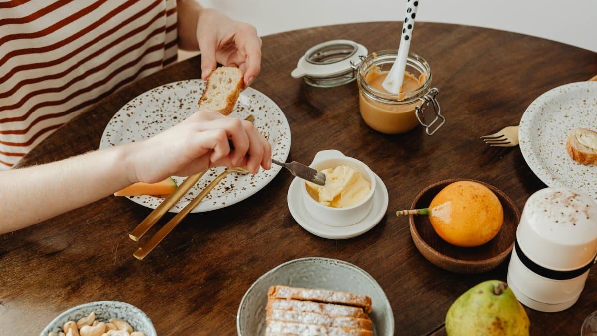 Overhead shot of a rustic breakfast scene with bread, butter, fruits, and nuts on a wooden table.