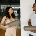 A smiling man and woman holding yoga mats, preparing for a fitness class in a modern gym.