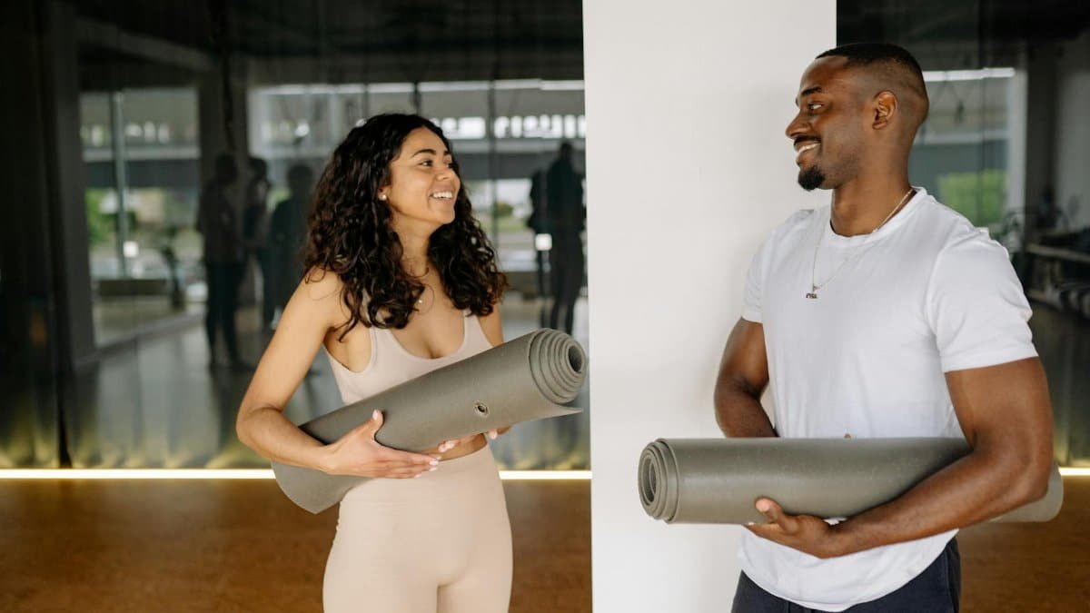 A smiling man and woman holding yoga mats, preparing for a fitness class in a modern gym.