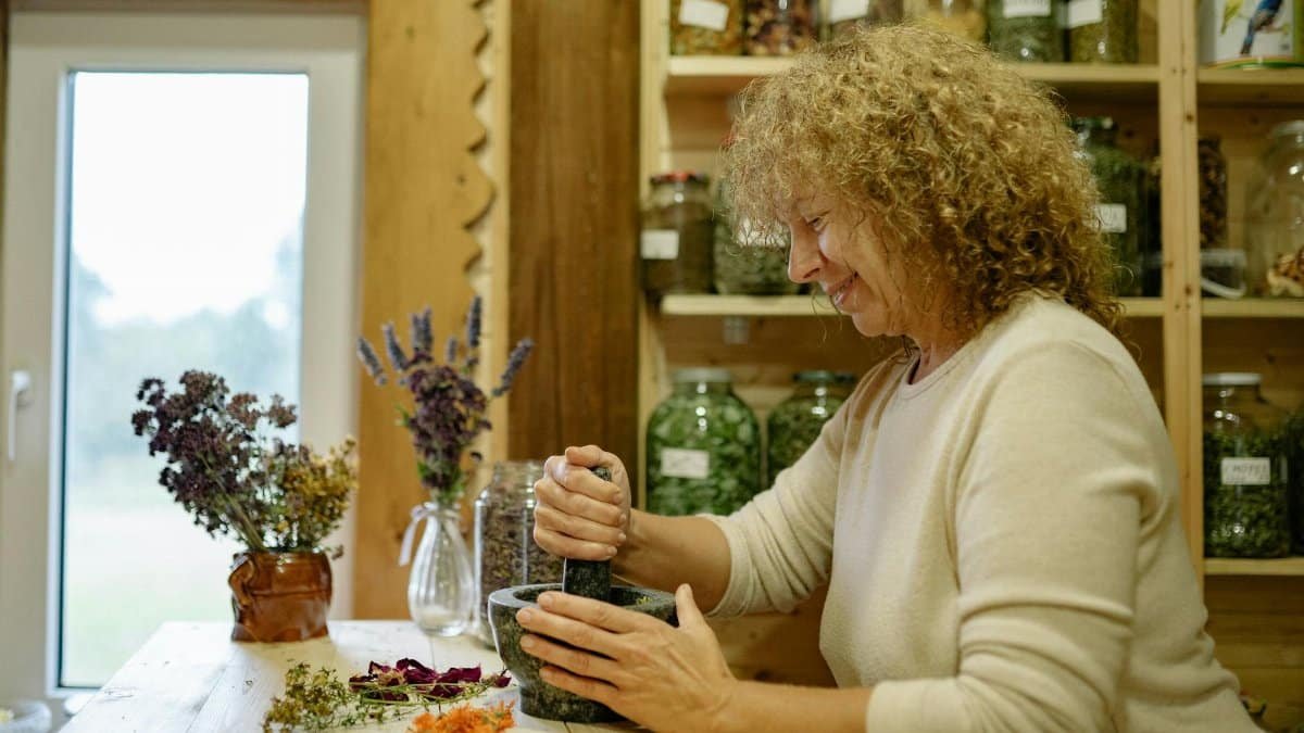 Woman grinding herbs in a mortar for natural remedies, surrounded by jars and plants.