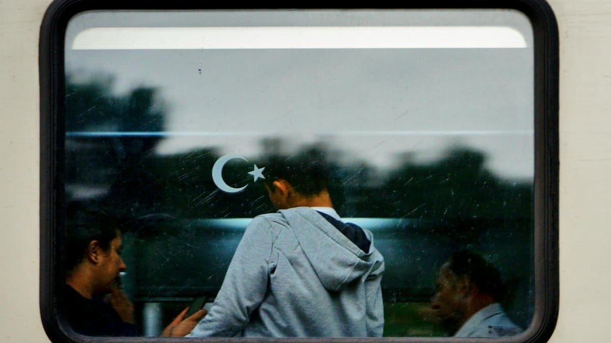 Three people captured through a train window, reflecting a scene with a crescent and star decal.