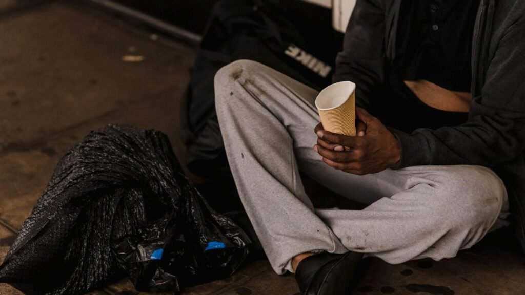Close-up of a man sitting on a city street holding a paper cup, highlighting urban life challenges.