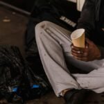 Close-up of a man sitting on a city street holding a paper cup, highlighting urban life challenges.