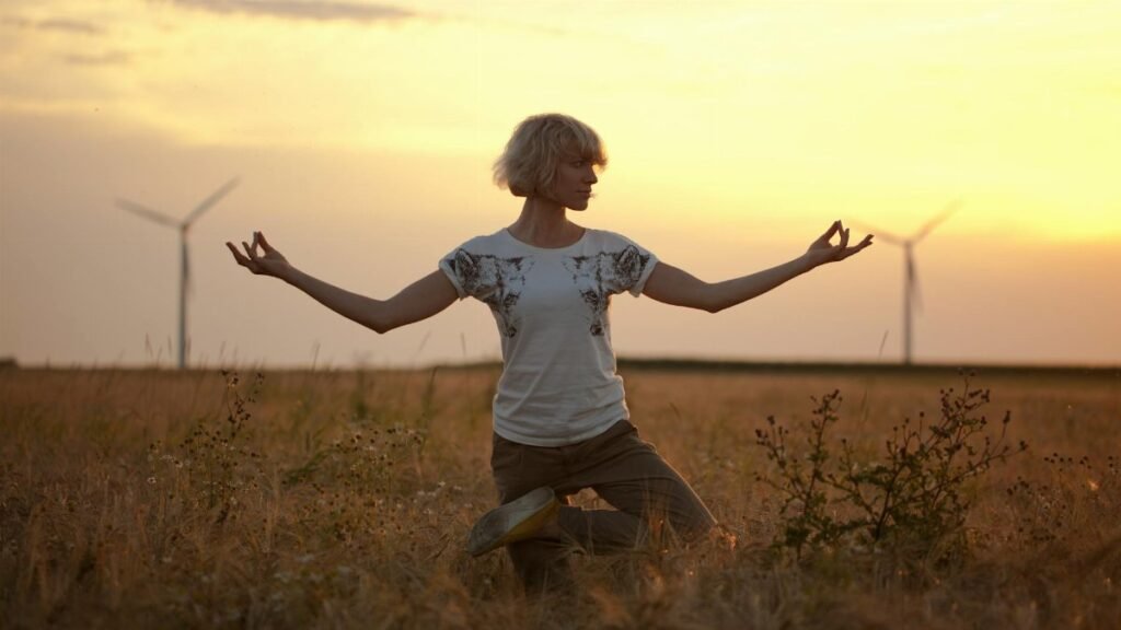 A woman practices yoga in a grassy field during sunset with wind turbines in the background.