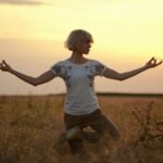 A woman practices yoga in a grassy field during sunset with wind turbines in the background.