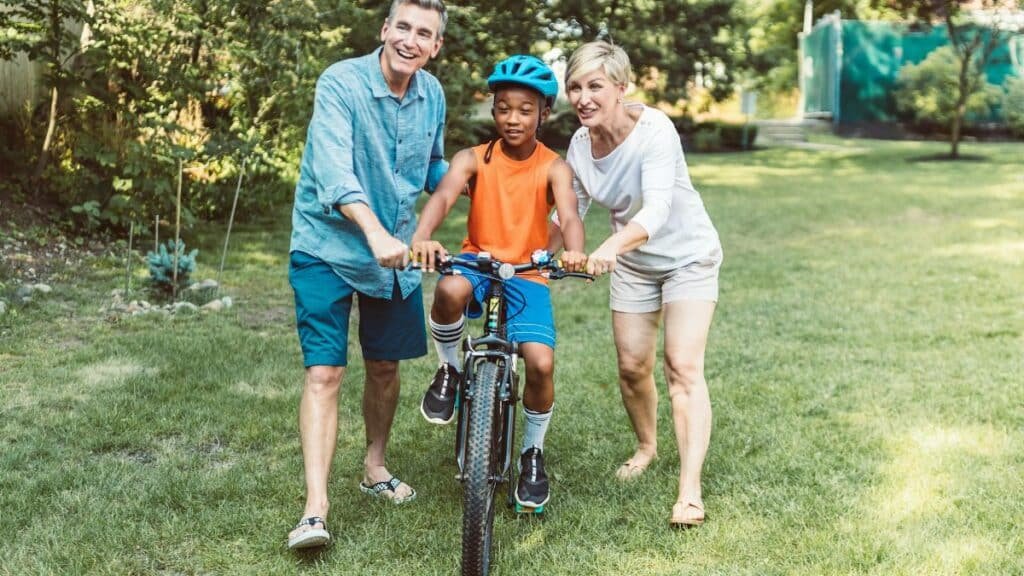 A family helps a young boy learn to ride a bicycle outdoors.