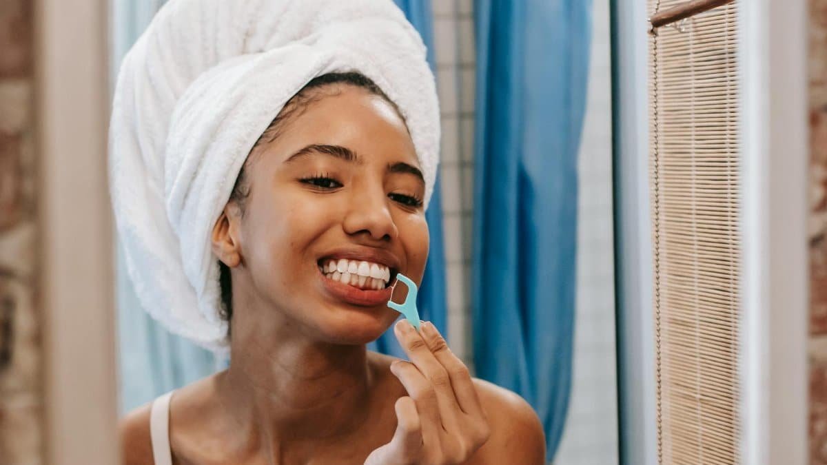 Young woman with towel on head flossing teeth in bathroom mirror, promoting hygiene and wellness.