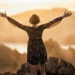 A woman stands with open arms on a mountain at sunset, embracing nature's beauty.