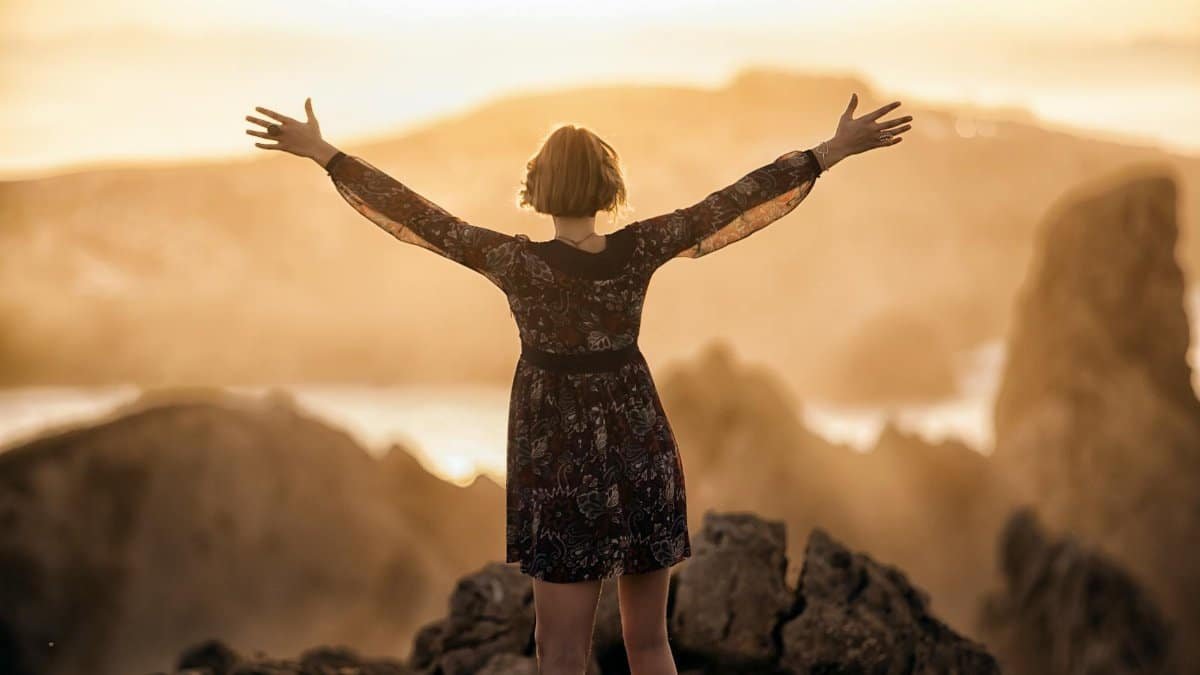 A woman stands with open arms on a mountain at sunset, embracing nature's beauty.