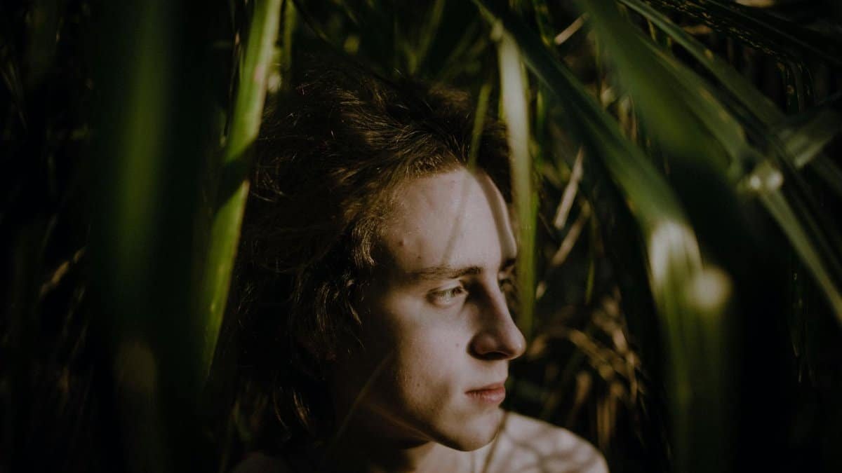 Young adult in thoughtful pose surrounded by lush green leaves, creating a serene nature scene.