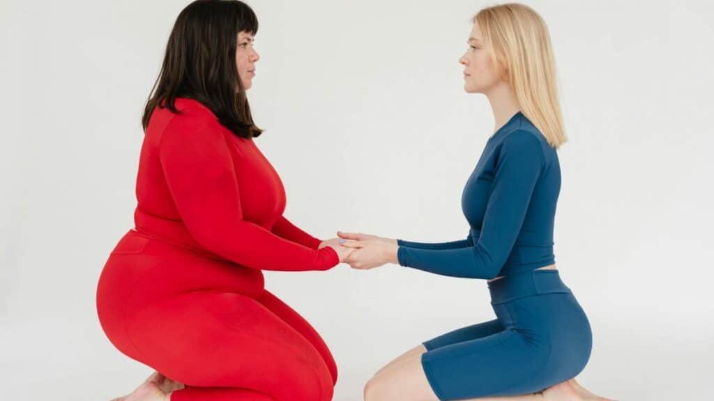 Full body side view of plus size woman and slender female instructor in activewear holding hands and looking at each other on white background