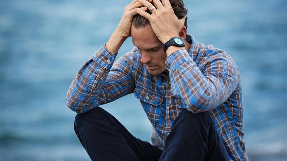 A man in a plaid shirt sits by the water looking distressed, symbolizing stress.