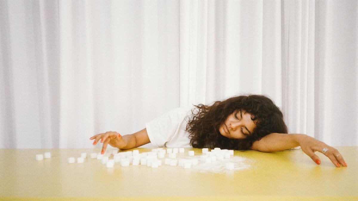 A woman rests her head on a table surrounded by sugar cubes, symbolizing fatigue or sugar crash.