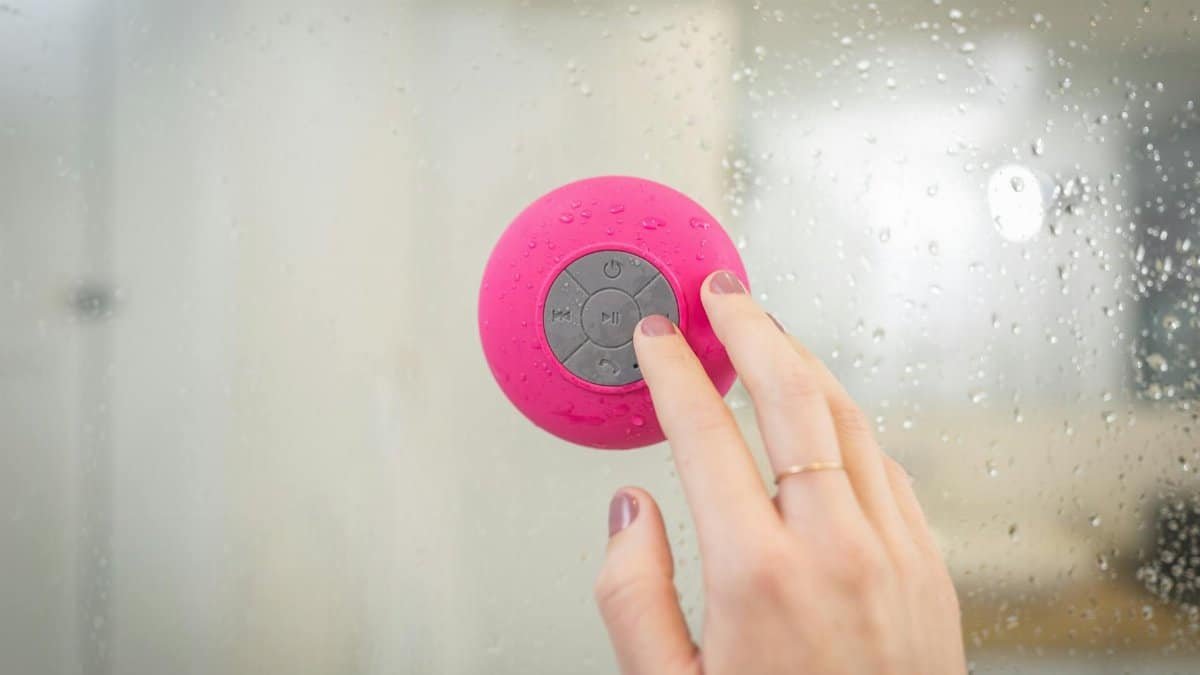 Close-up of a hand operating a waterproof speaker in a steamy shower environment.