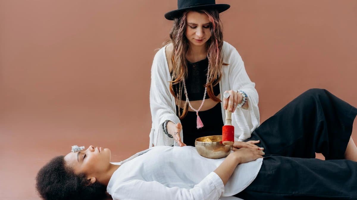 Woman practicing sound healing with singing bowl over another woman. Meditative and relaxing wellness session.