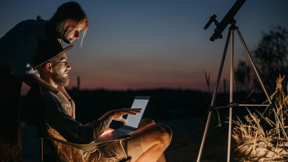 A couple using a laptop and telescope under the night sky, enjoying stargazing outdoors.
