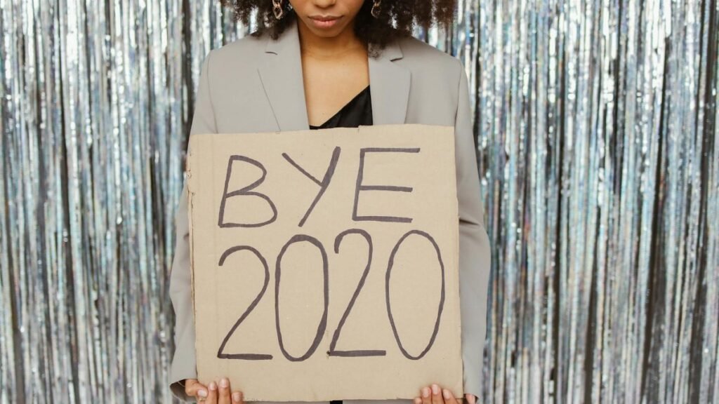 Young woman holding a 'Bye 2020' sign in front of a shiny, reflective backdrop, expressing relief and new beginnings.