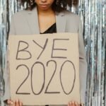 Young woman holding a 'Bye 2020' sign in front of a shiny, reflective backdrop, expressing relief and new beginnings.