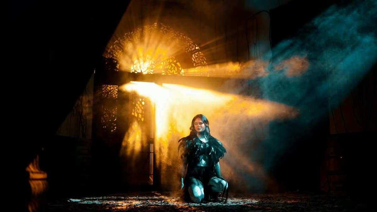 A young woman kneels in dramatic light and shadows, creating an ethereal atmosphere.