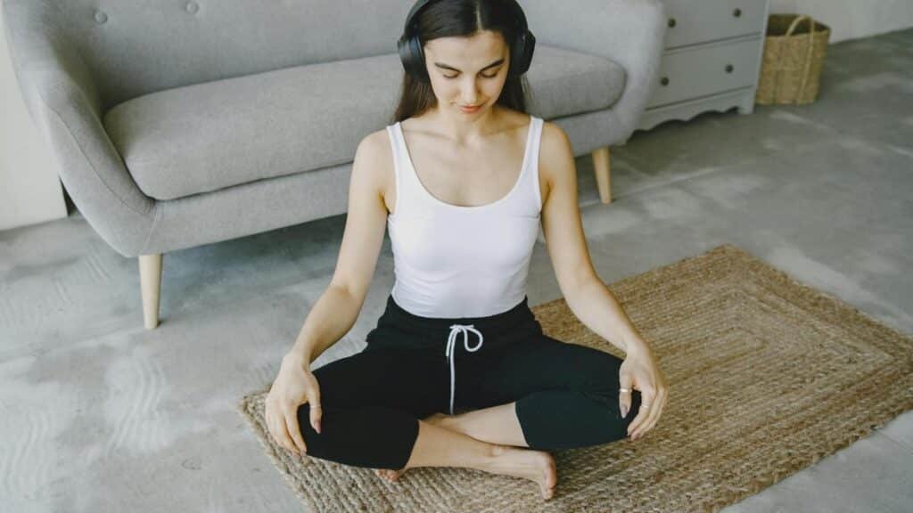 Woman meditating indoors on a rug with headphones, promoting relaxation and wellness.