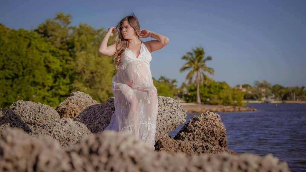 Pregnant woman in lace dress posing on rocks by Miami shoreline amidst lush greenery.