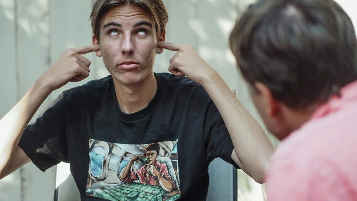 A teenage boy in a black shirt covers his ears and looks up, ignoring a conversation outdoors.