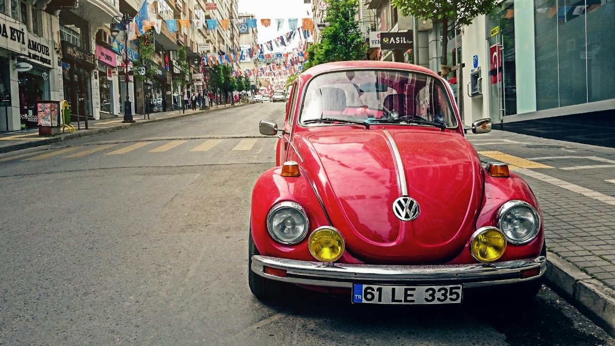 Bright red vintage Volkswagen Beetle parked on a colorful urban street with flags overhead.