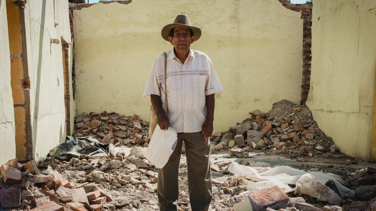 An elderly man stands in debris of a demolished building, showcasing resilience after disaster.