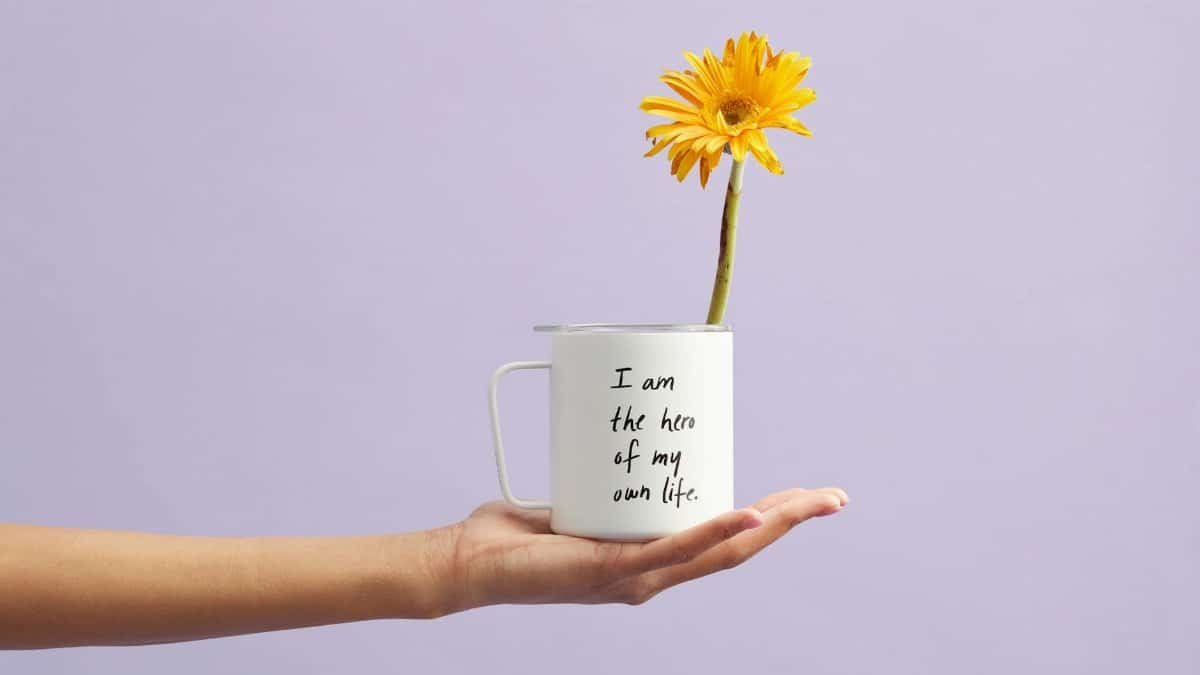 Hand holding a mug with a sunflower and motivational quote, set against a purple background.