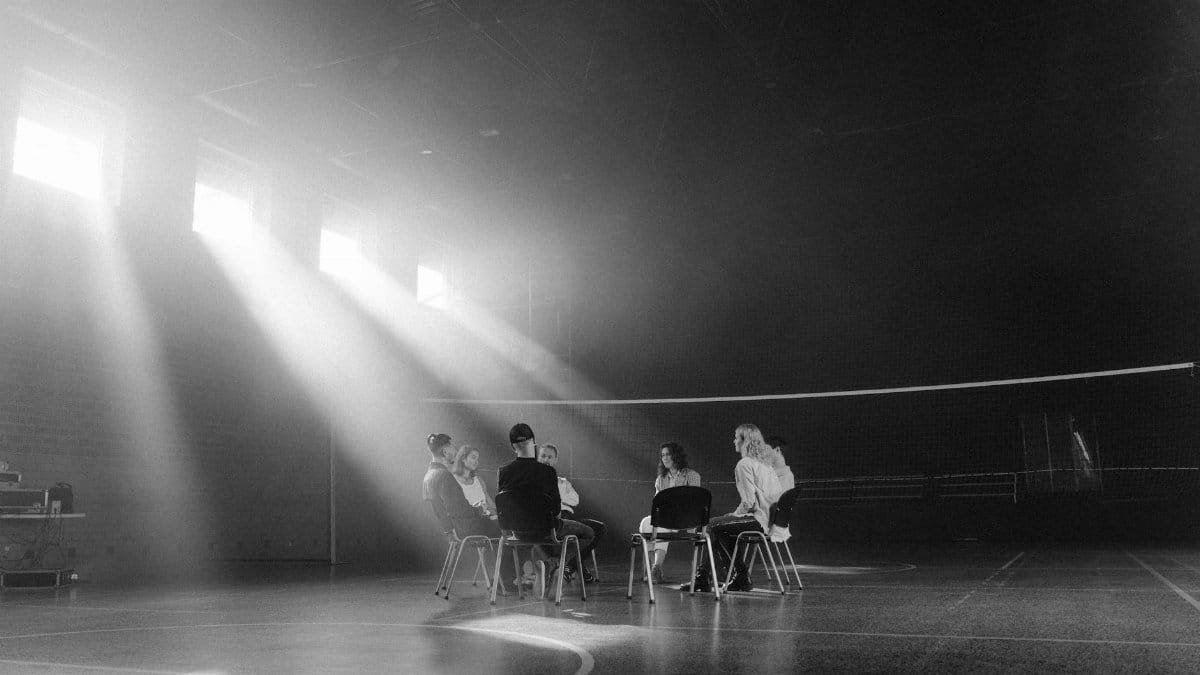 A group of people having a discussion in a sunlit indoor sports hall.