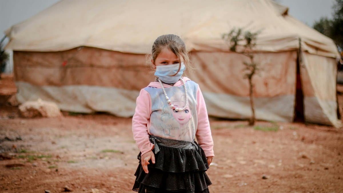 A girl wearing a face mask stands outside a tent in a refugee camp in Syria.