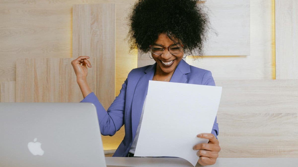 A cheerful woman celebrates her success at work, looking at a document in an office setting.