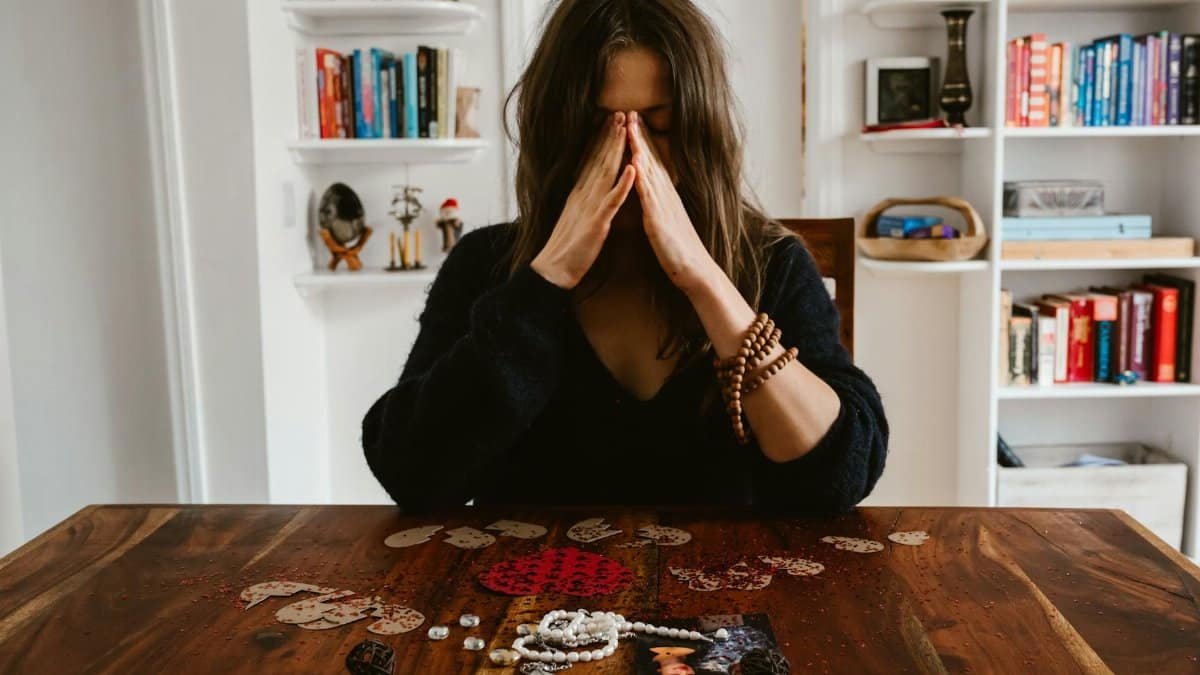 A woman in deep thought sits at a table with beads and a photo, conveying introspection.