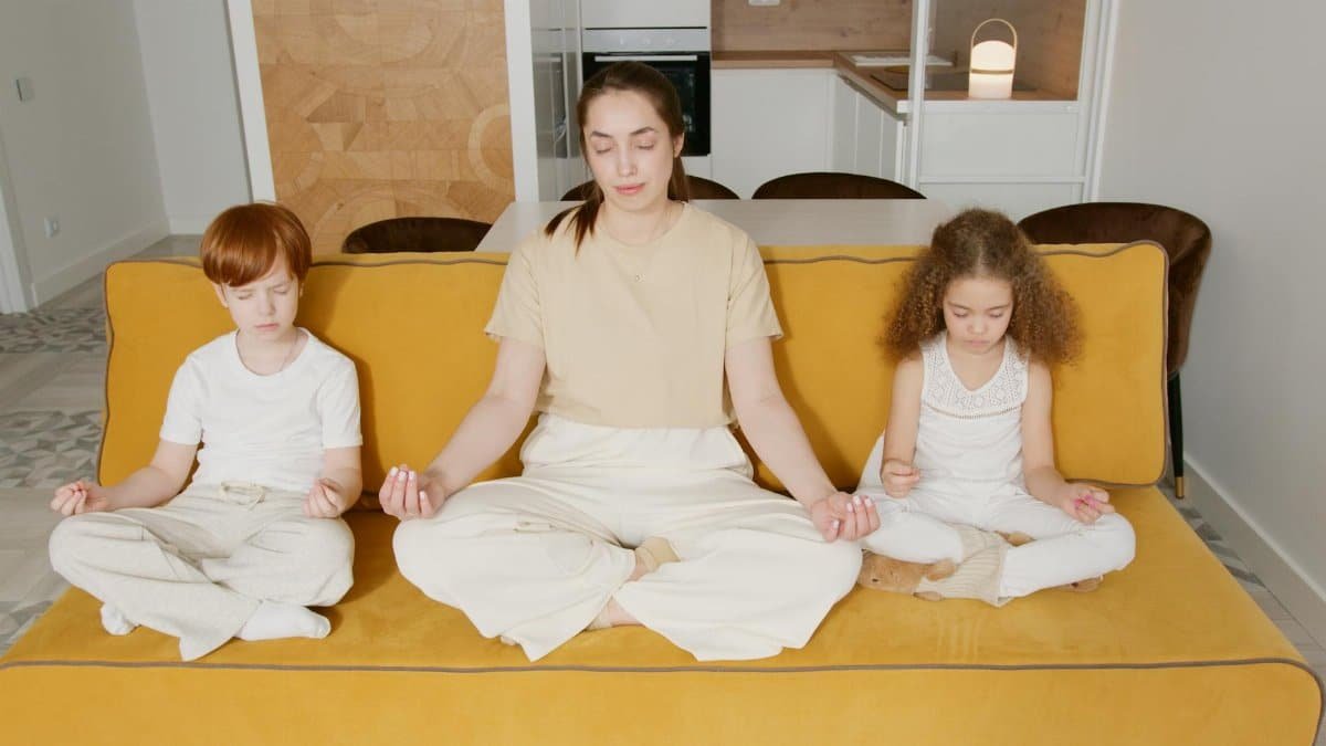 A mother and two children practicing mindfulness meditation indoors on a cozy sofa.