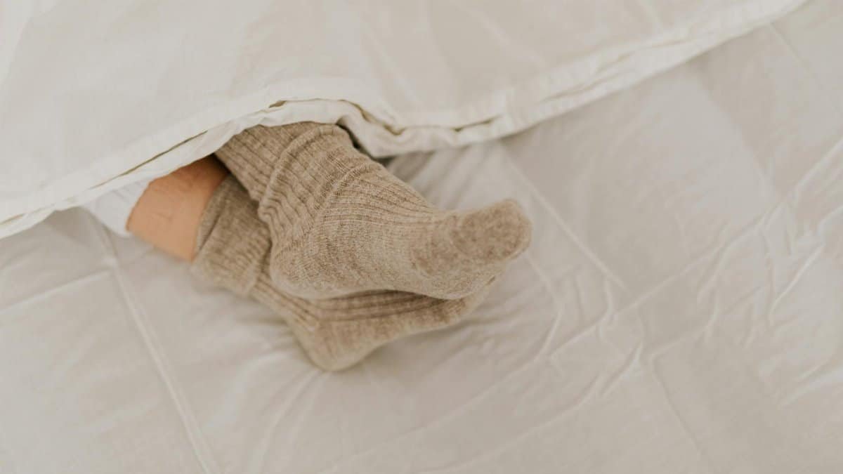 Comfortable feet in woolen socks peek out under a soft white linen blanket on a bed.