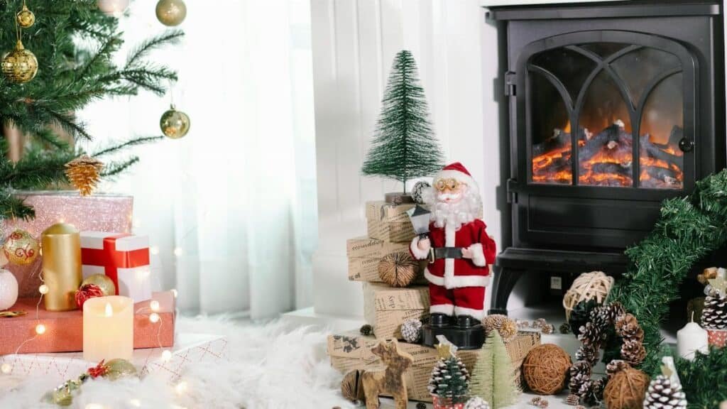 Cozy light room with gift boxes under Christmas tree and Santa Claus near holiday decorations placed on white cozy fabric at home with fireplace and burning candles