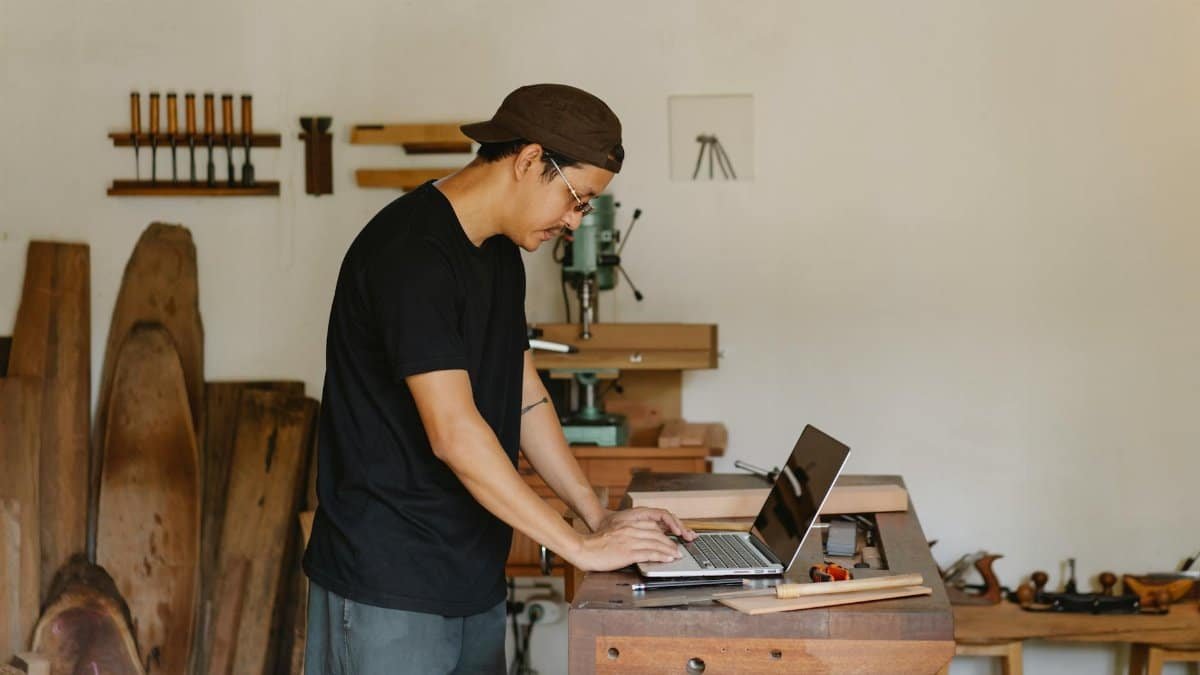 A craftsman focuses on his laptop in a woodworking workshop, blending technology and traditional craft.