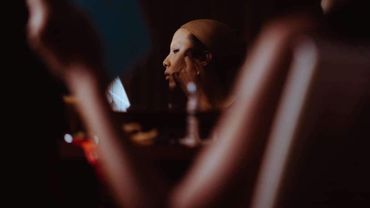 Asian woman applying makeup with brush in dimly lit room, focused on her reflection.