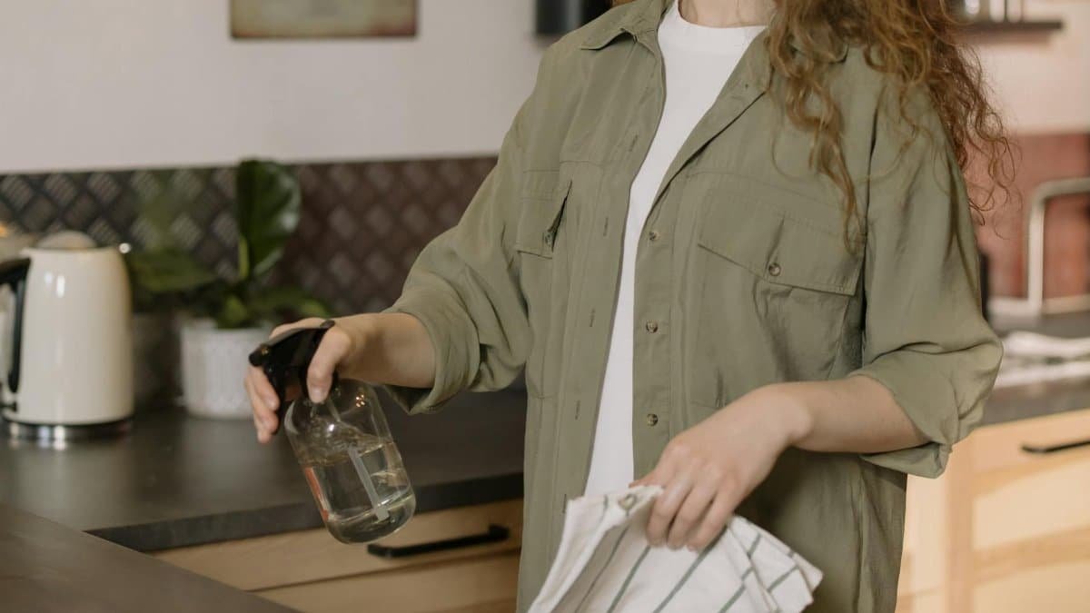 A woman with curly hair cleans a kitchen counter using a spray bottle and cloth.